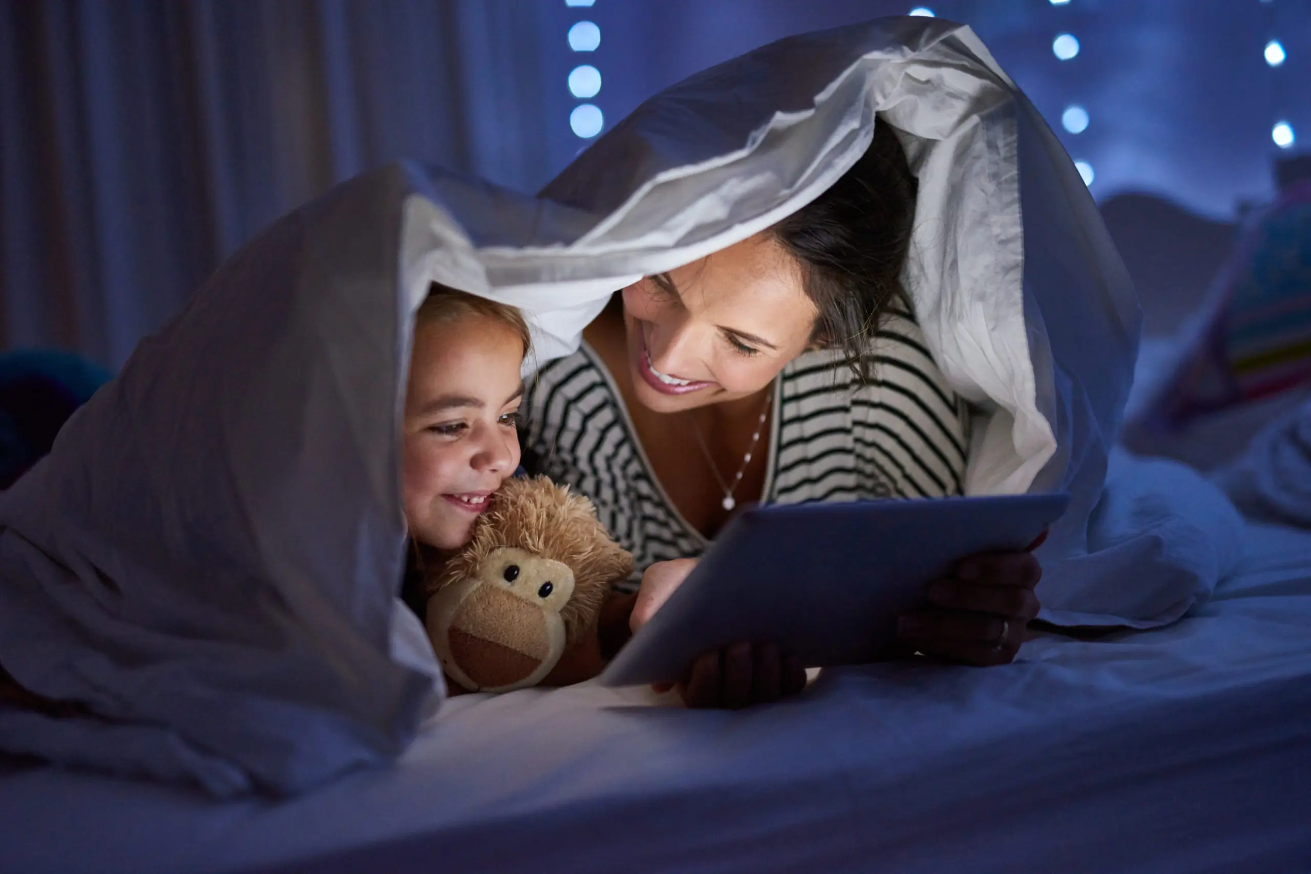 Bild einer Mutter und ihrer Tochter, welche gemeinsam unter der Bettdecke liegen und mit einem Tablet interagieren